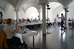 People sit at a long table with a white tablecloth. Some raise their hands.